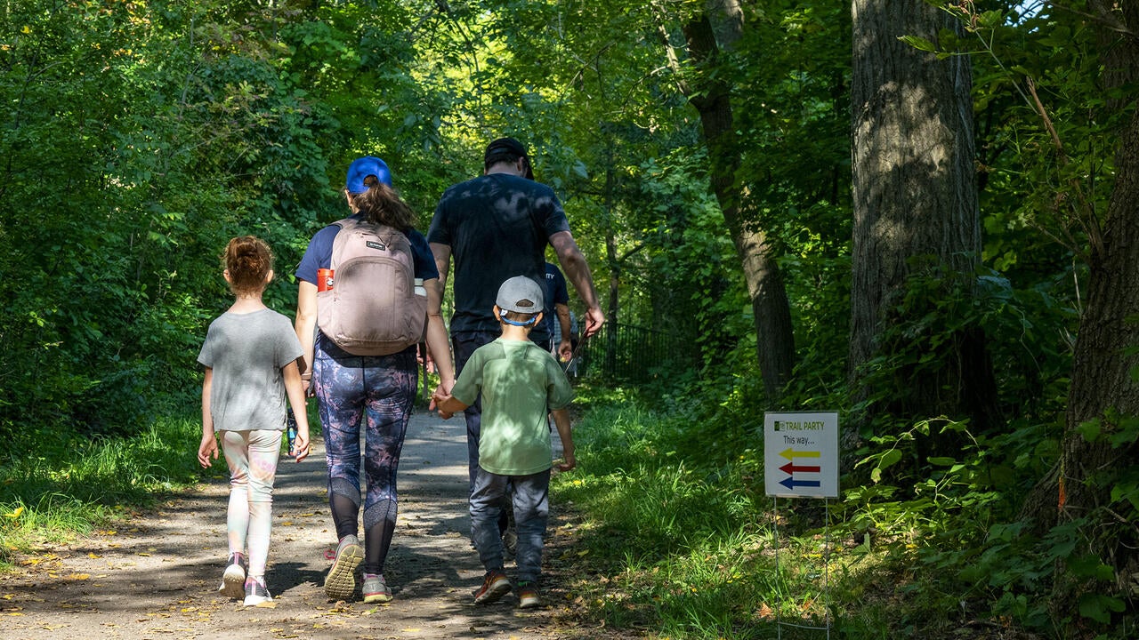 A family of two adults and two small children walk away from the camera down a woodland path