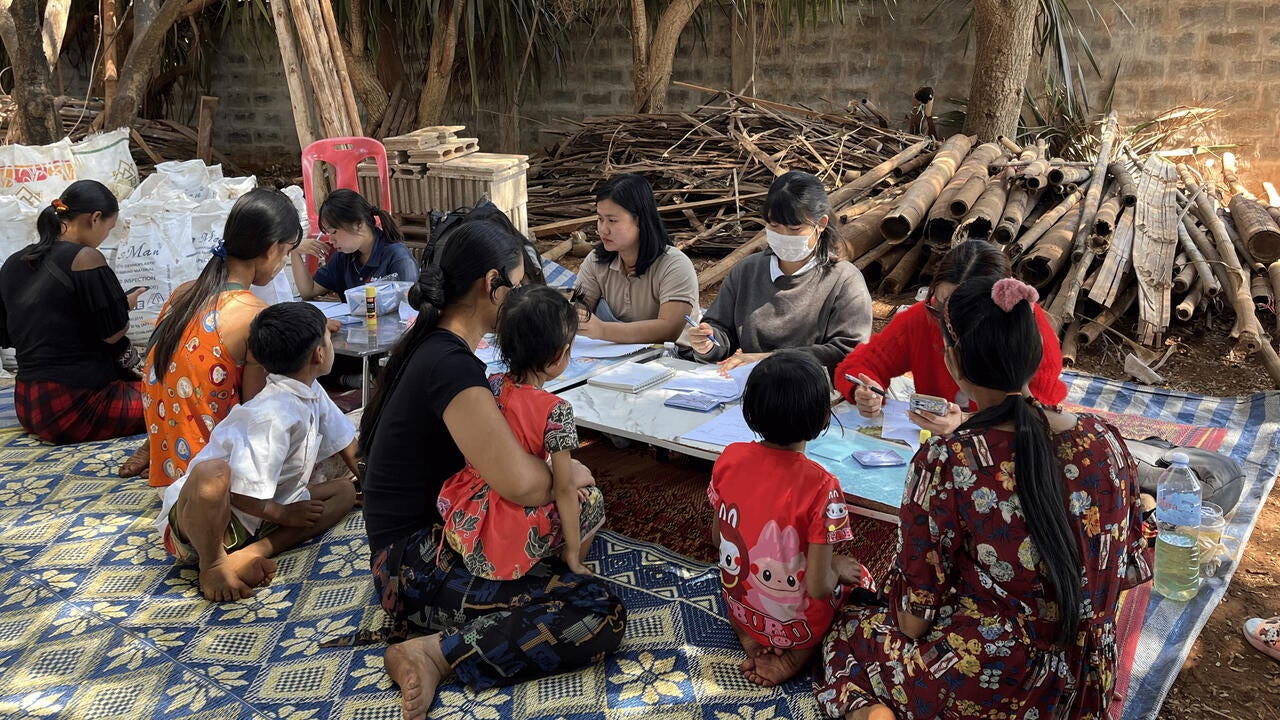 A group of women and children sit outdoors around a low table covered with papers and medical supplies.